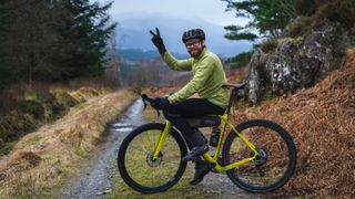 A gravel rider gives the peace sign with cloud obscured mountains in the background