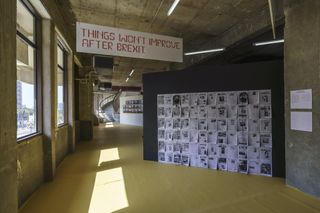 Some graphic design artworks are showcased inside the building. We see a sign hanging from the wall that says 'Things won't improve after Brexit'. There are many, what looks like, newspaper pages hung on a black platform.