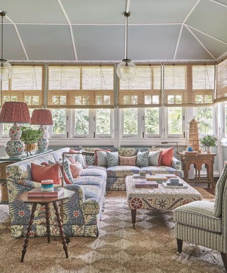 Maximalist living room with large l-shaped patterned sofa with matching ottoman, two large lampshades on console, jute rug, wicker-style blinds on large windows