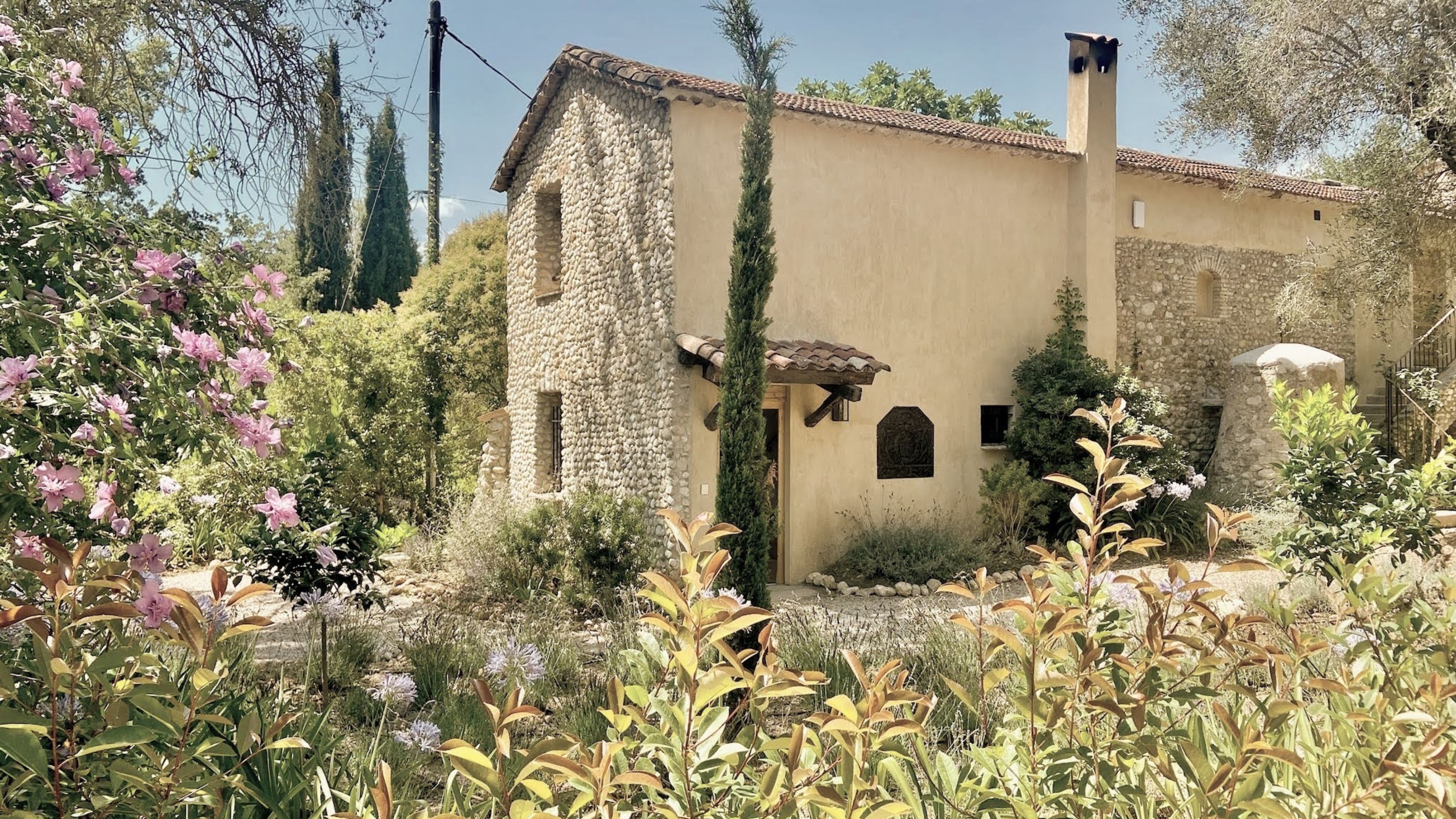 A rustic stone and stucco villa at Toile Blanche featuring a traditional tiled roof, surrounded by lush Mediterranean gardens and vibrant pink bougainvillea.