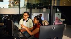 Male and female cybersecurity workers talking while looking at desktop computer screen in an open plan office space, with colleagues relaxing in background.