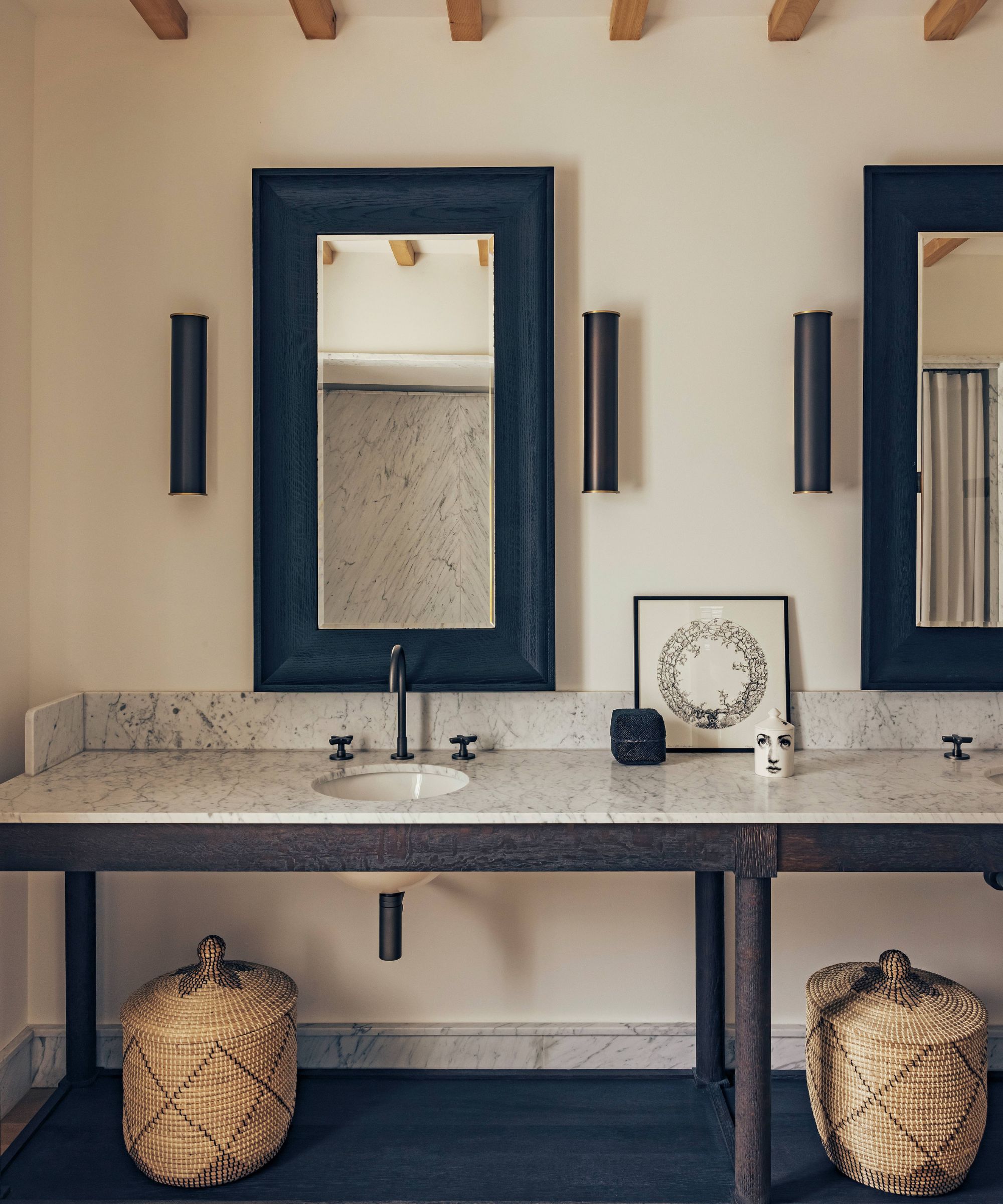 a neutral rustic bathroom in a home in france with ceiling beams, black wooden and marble double vanity and rattan baskets