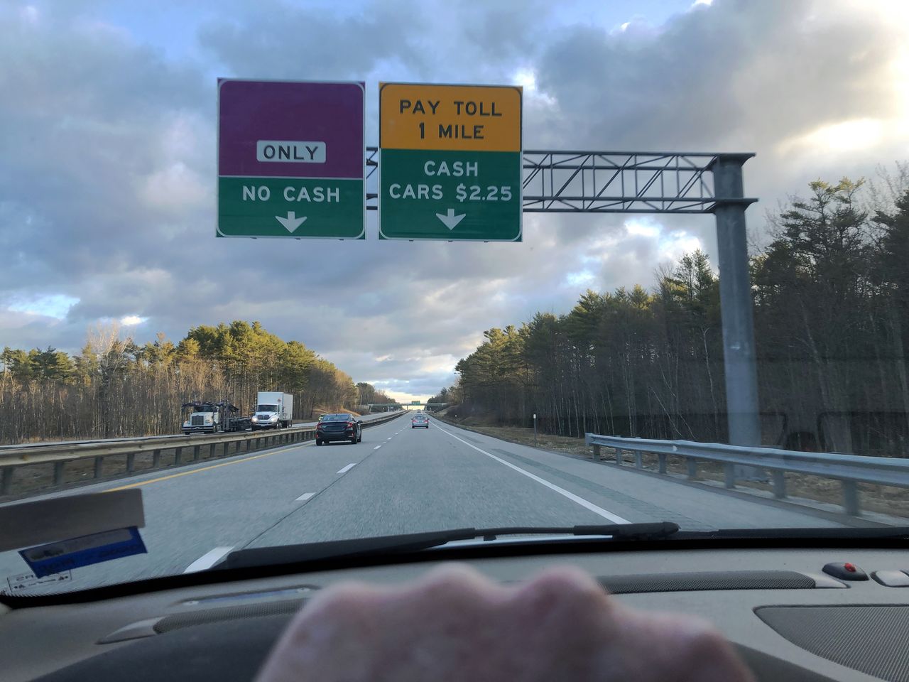 The top of a hand on a steering wheel, and past the car window a highway and a sign for an upcoming toll.