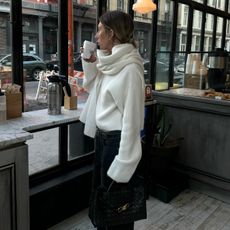style influencer and stylist Hannah Lewis poses while drinking coffee in a café wearing a chic winter 2026 outfit with a cream scarf sweater dark jeans and a Bottega Veneta bag