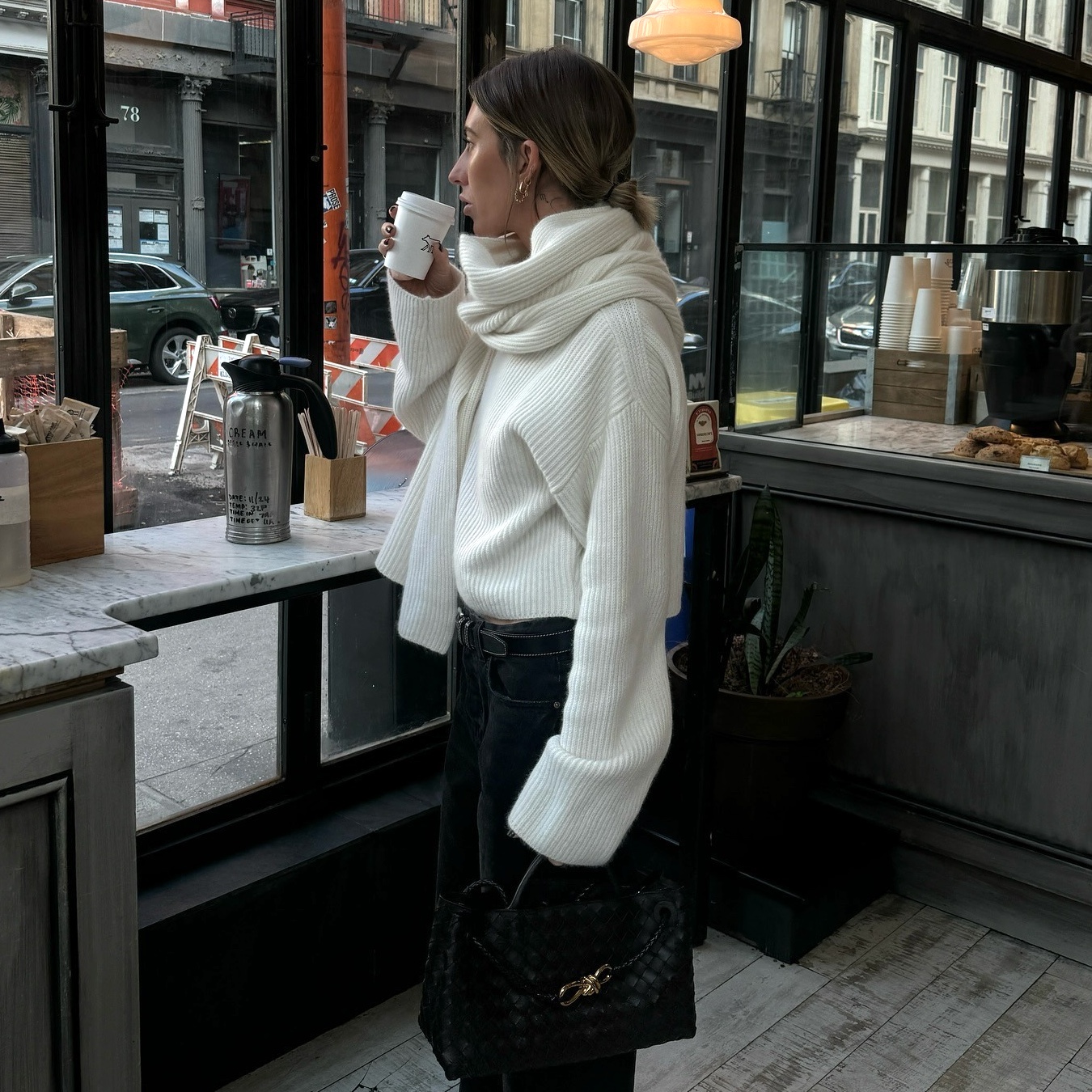 style influencer and stylist Hannah Lewis poses while drinking coffee in a café wearing a chic winter 2026 outfit with a cream scarf sweater dark jeans and a Bottega Veneta bag