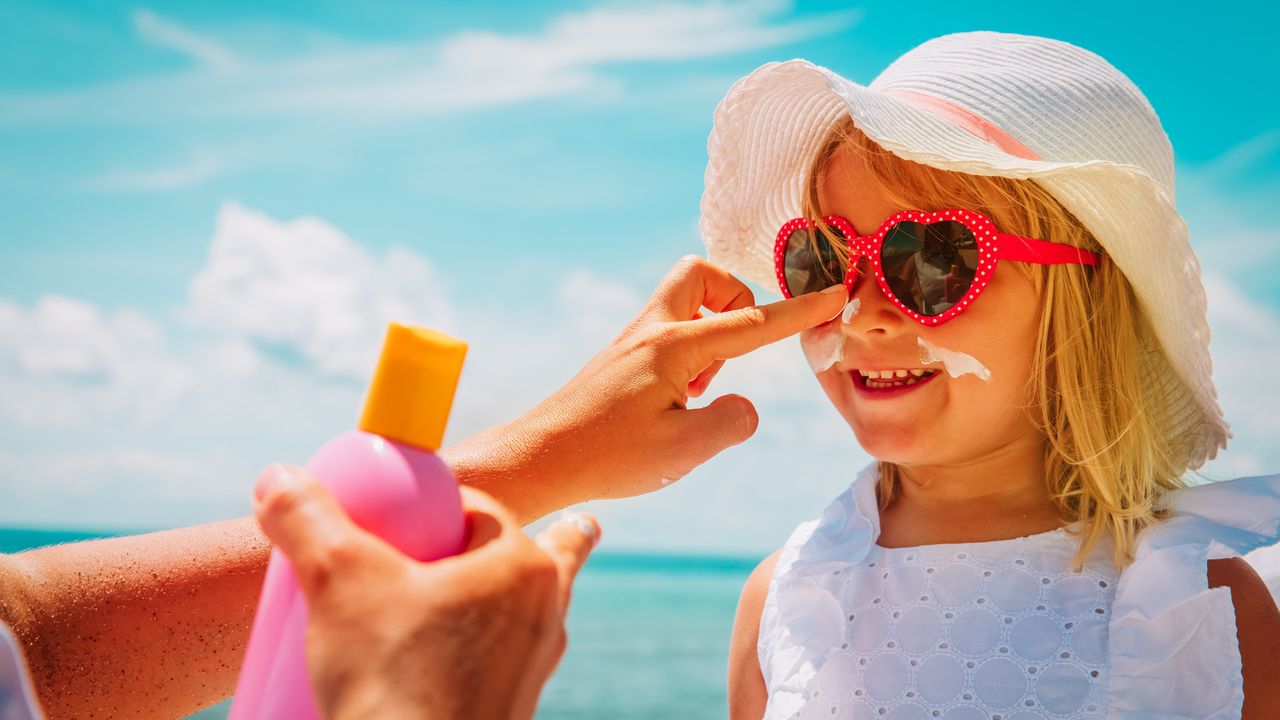 best sunscreen for children - image of a small girl wearing sunglasses and a sun hat having sun cream applied to her facechildren