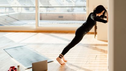 A woman performs push-ups on a wall, with her hands against its surface and her feet stepped back. Behind her is a yoga mat, a water bottle, a resistance band and a laptop.