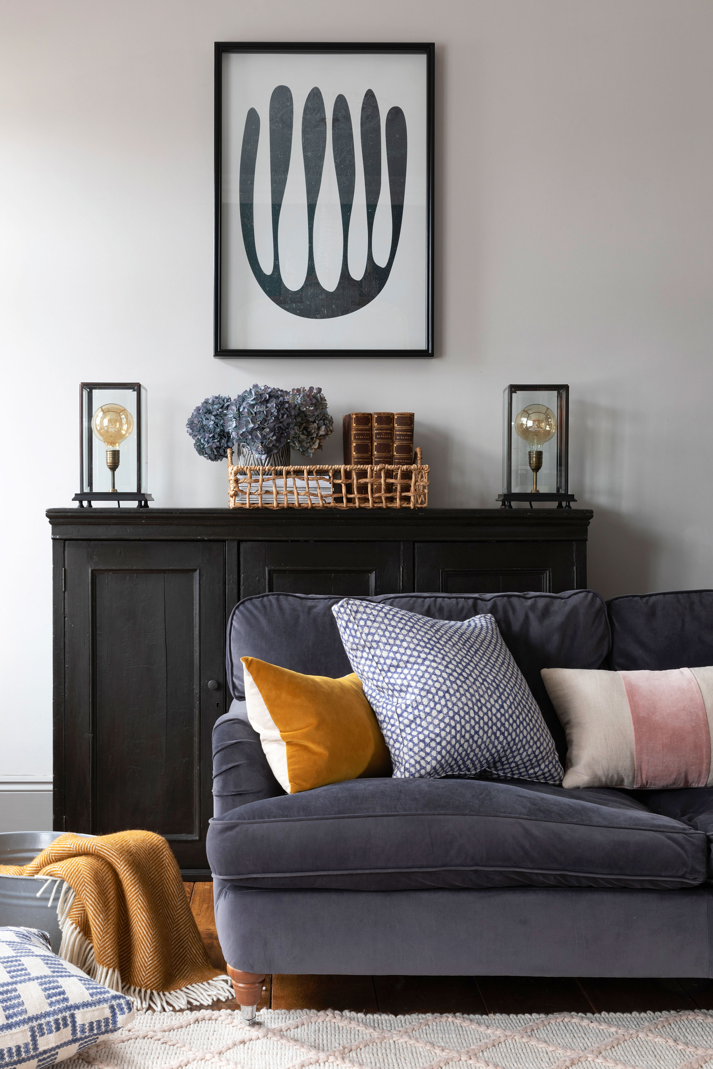 A grey living room with dark grey sofa, black living room cabinetry storage, monochrome abstract wall art, woven basket, Edison-style decorative lights and mustard, blue and pink cushions on sofa