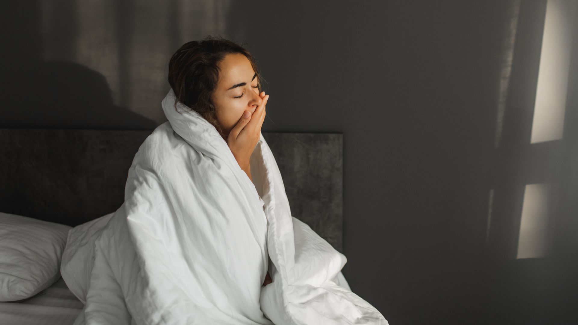 A woman sits up in bed covering her mouth with her hand as she yawns. She has her white duvet wrapped around her shoulders.
