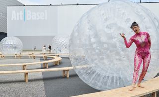 An art exhibit with round wooden walkways with female models walking on them and large round clear plastic balls rolling between them.