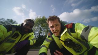 A point of view image from Flynn's perspective of paramedics Jacob and Iain in their uniforms looking at him with concern by the side of a road.