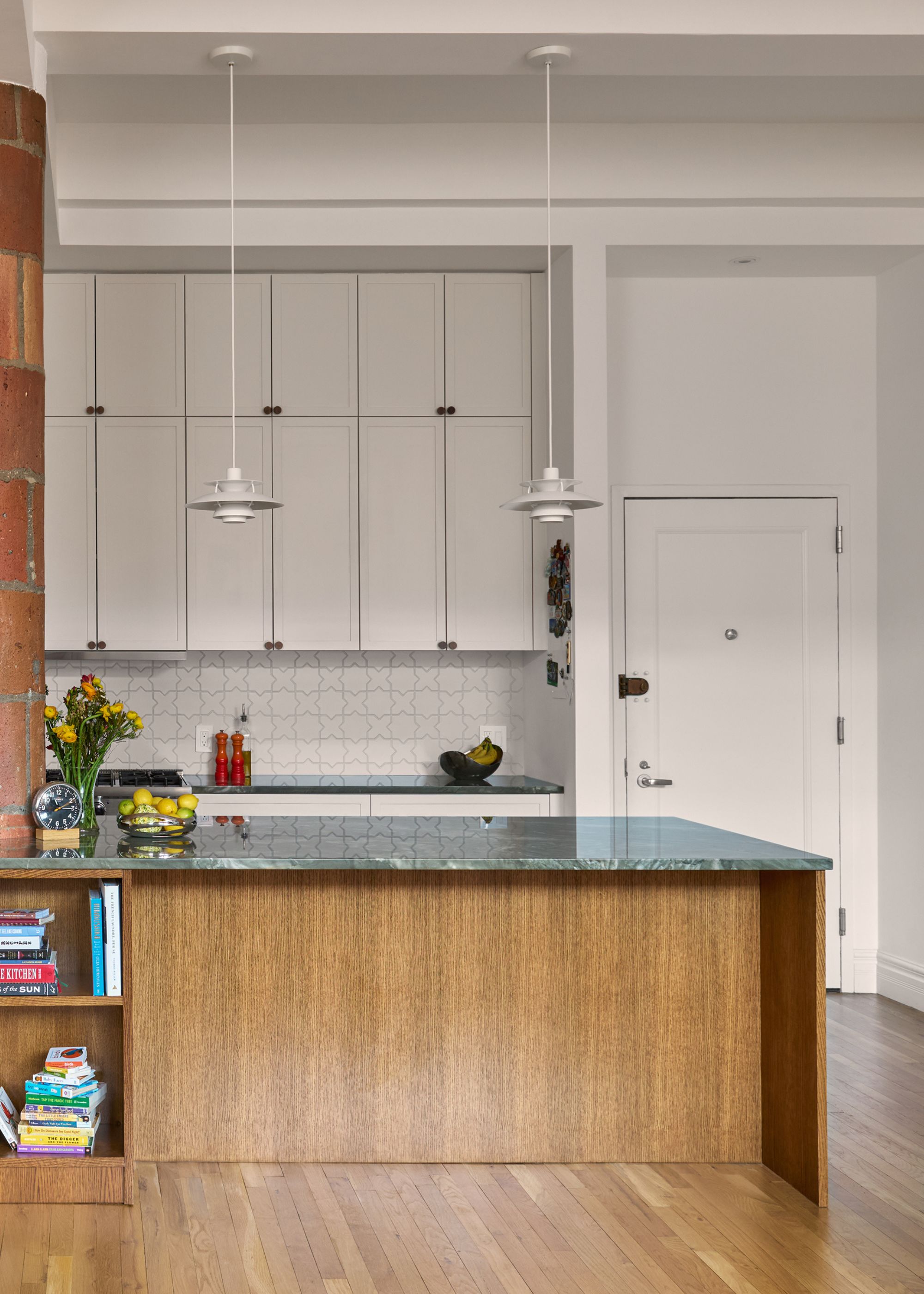An apartment kitchen with light gray walls and built-ins, a wooden island with dark green marble countertops, wood flooring, and a tall ceiling with two white modern pendant lights