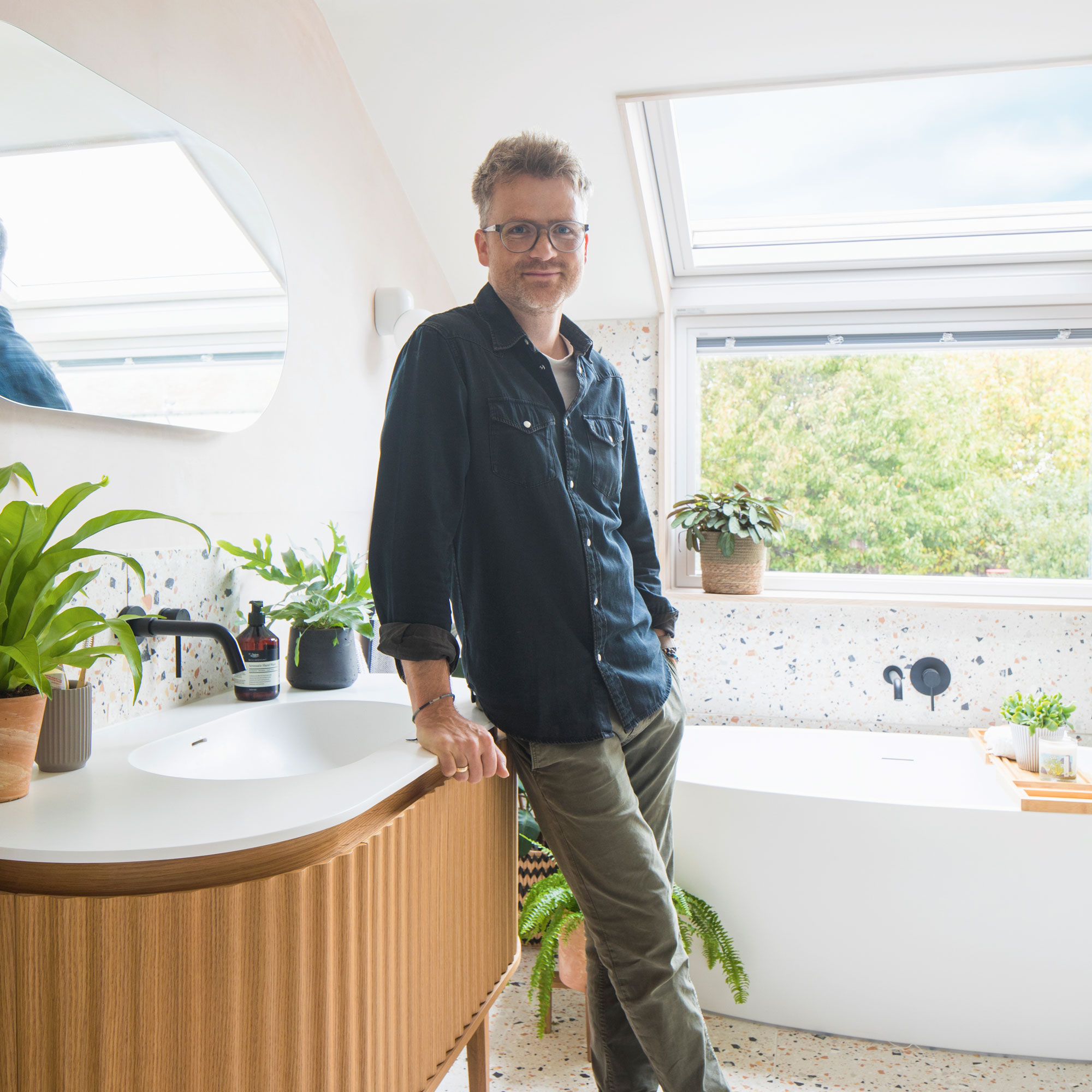 Bathroom with terrazzo on wall and floor, a fluted wooden vanity with white countertop, a freestanding bathtub, pink wall with lozenge shaped mirror and a wall mounted light. A man is leaning on the vanity.