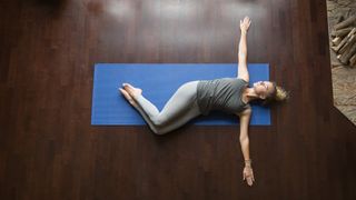 A woman lies on her back on a yoga mat and performs a supine twist. Her arms lie out to either side of her, in line with her shoulders. Her legs are bent gently at the knee and flopped over to her left side.