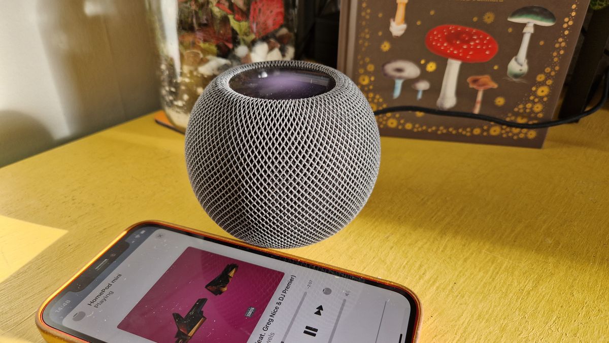 A dark grey Apple HomePod Mini smart speaker on a piece of yellow furniture. There is an iPhone in the foreground showing some music playing.