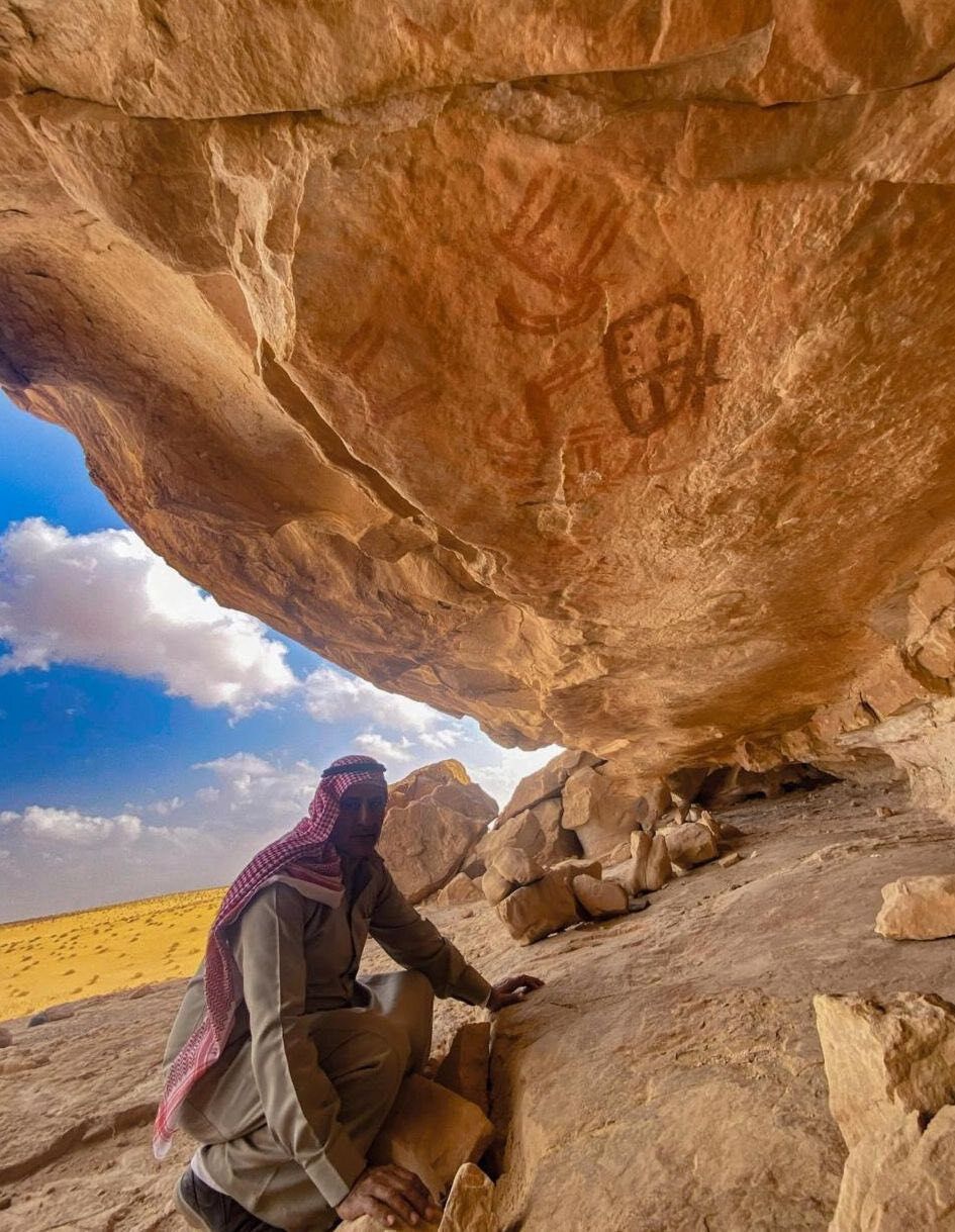 A man wearing a headscarf leans underneath a sheer reddish rock face with large dark inscriptions on it.