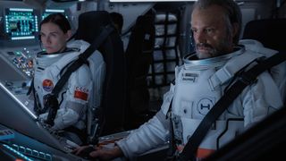 A woman and a man wearing white astronaut suits inside a spaceship cockpit.
