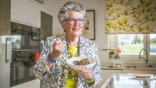 Prue in her kitchen smiling for the camera and wearing a bright top and necklace and holding a fork and bowl as if she is about to eat 