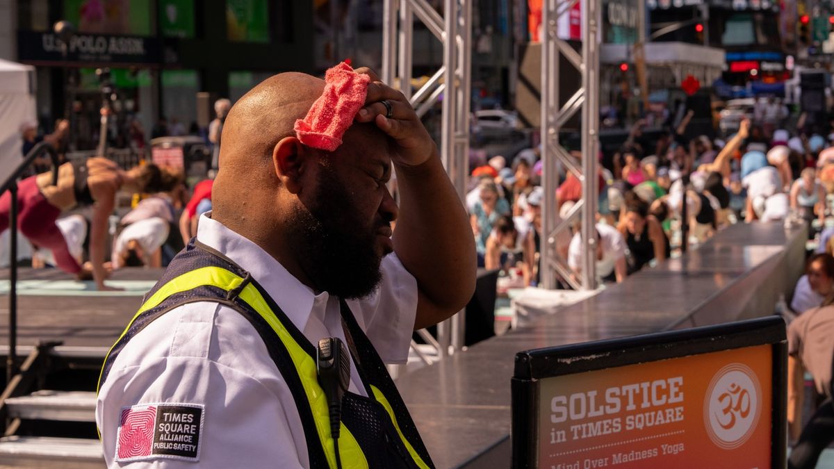 A public safety officer in a yellow high-vis jacket and white shirt is shown sweltering in the heat. He is holding a red cloth to this forehead to wipe away sweat. 