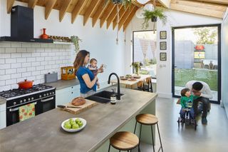 A kitchen with large pivot door and picture window, concrete-effect worktops and floor, and exposed beams in the pitched extension roof