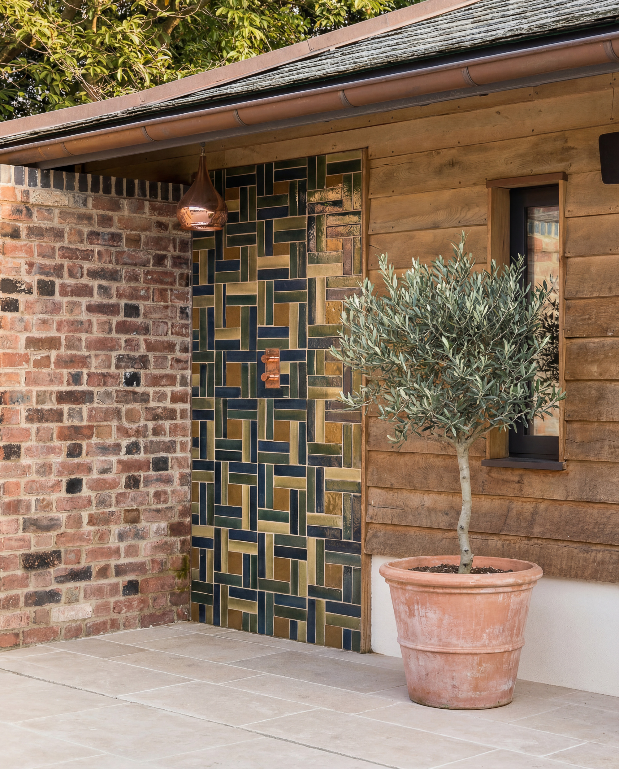 outdoor shower with decorative Ca' Pietra tiles, a brick wall, timber cladding, a potted olive tree, and a Maroc Khaki Exterior Shower head
