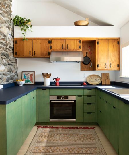 Rustic U-shaped kitchen with vibrant green lower cabinets, dark blue countertops, and vintage-style honey wood upper cabinets next to a stone wall.
