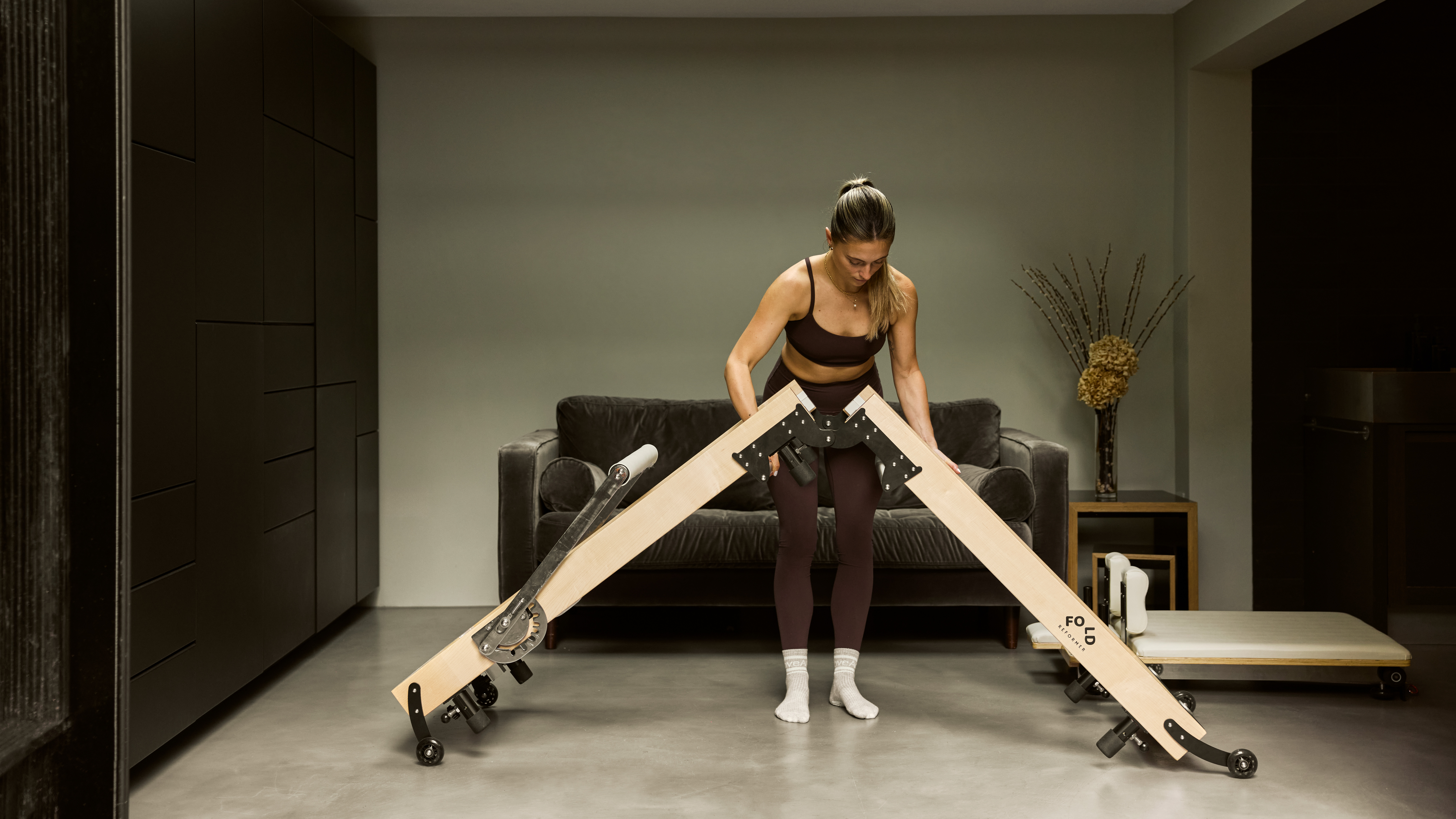 FOLD Reformer bed being assembled and extended by a woman in Pilates workout gear at home