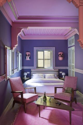 A hotel bedroom with a bed, triangular side tables with lamps, two pink chairs, a triangular rounded coffee table, a ping rug, purple walls and a large widow with shutters.