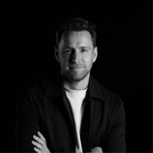 Black-and-white image of a man in a white shirt and black shacket standing in front of a dark background.