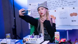 A content creator (Sia Jiwoo) takes a selfie in a streaming room with a whiteboard, on Netflix's 'The Influencer.'