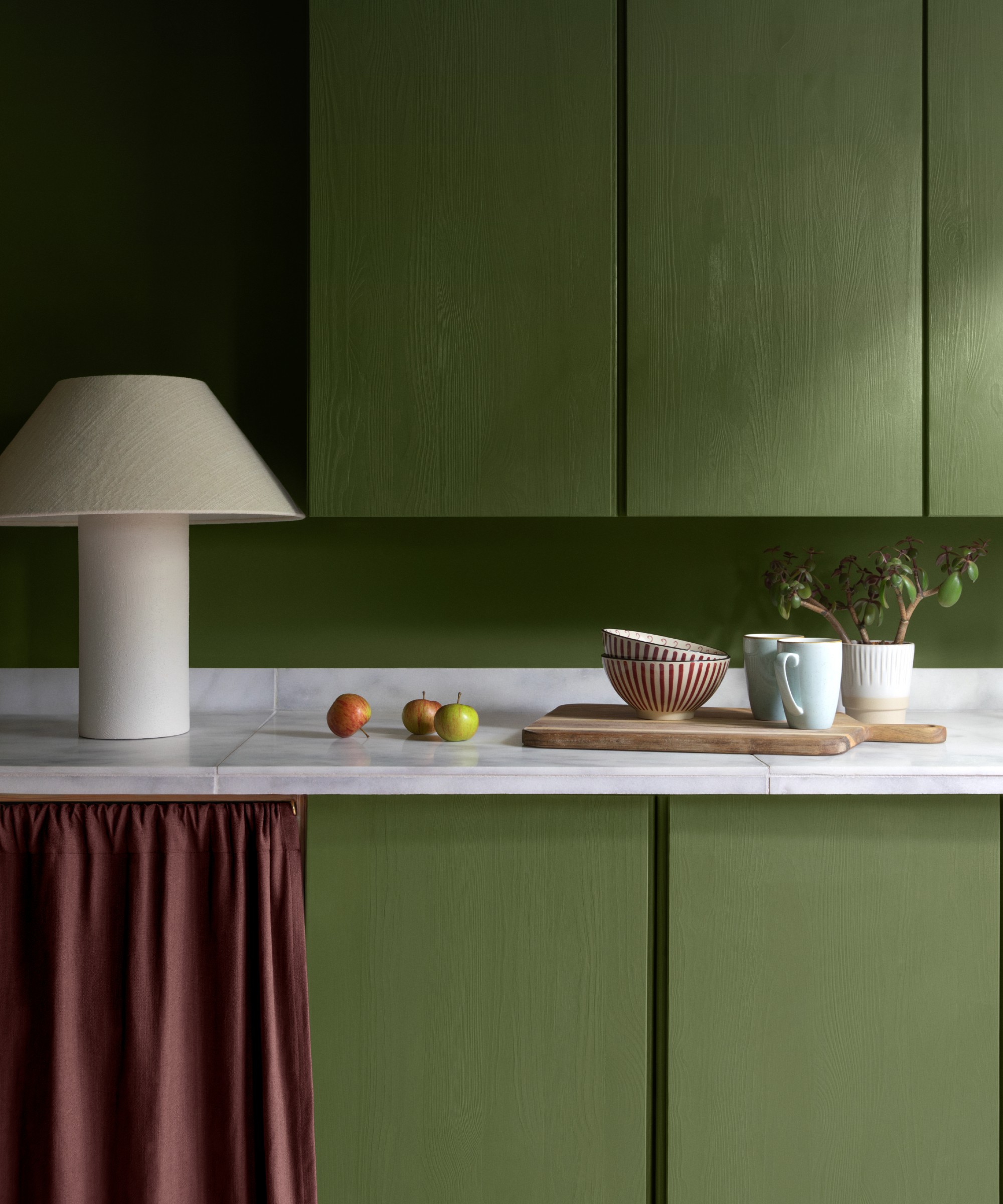 A green kitchen painted in Lick&#039;s Green 18 shade with a wine red curtain and an off-white table lamp