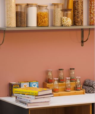 A pink kitchen with wall shelves with pantry jars and a countertop with spices, shelving, and books