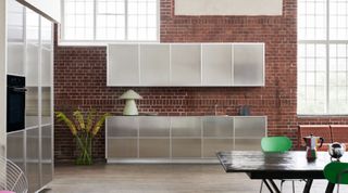 Silver, semi-transparent kitchen units, dark wood dining table and plant in a clear vase against a brick wall