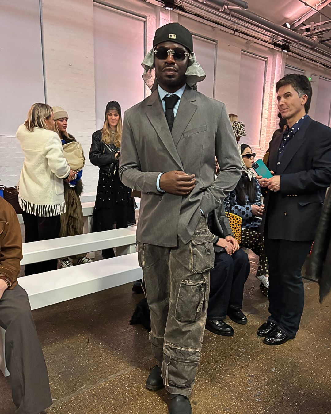 an image of Victoro, a fashion influencer (@victorolusina) at London Fashion Week, wearing grey denim wash cargo style trousers and a grey blazer, with glasses and a cap