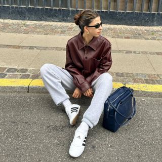 Daniela wearing Adidas Taekwondo white sneakers with grey sweatpants, a burgundy leather jacket, and a navy handbag.