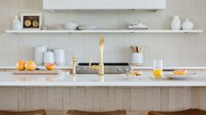 Learning how to brighten a small kitchen will achieve airy looks like this white kitchen with white shelves, a countertop with oranges on it and a tap, plus three wooden stools