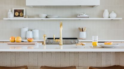 Learning how to brighten a small kitchen will achieve airy looks like this white kitchen with white shelves, a countertop with oranges on it and a tap, plus three wooden stools