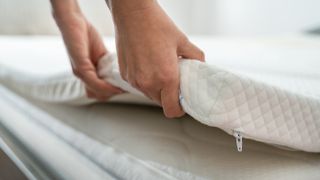 A person lifts a white mattress topper off a bed to check if it needs replacing as mattress toppers only last a few years