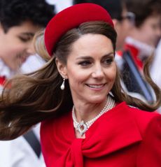 Kate Middleton wearing a red coat with a bow and a matching hat smiling as her hair blows in the breeze at Commonwealth Day 2025 