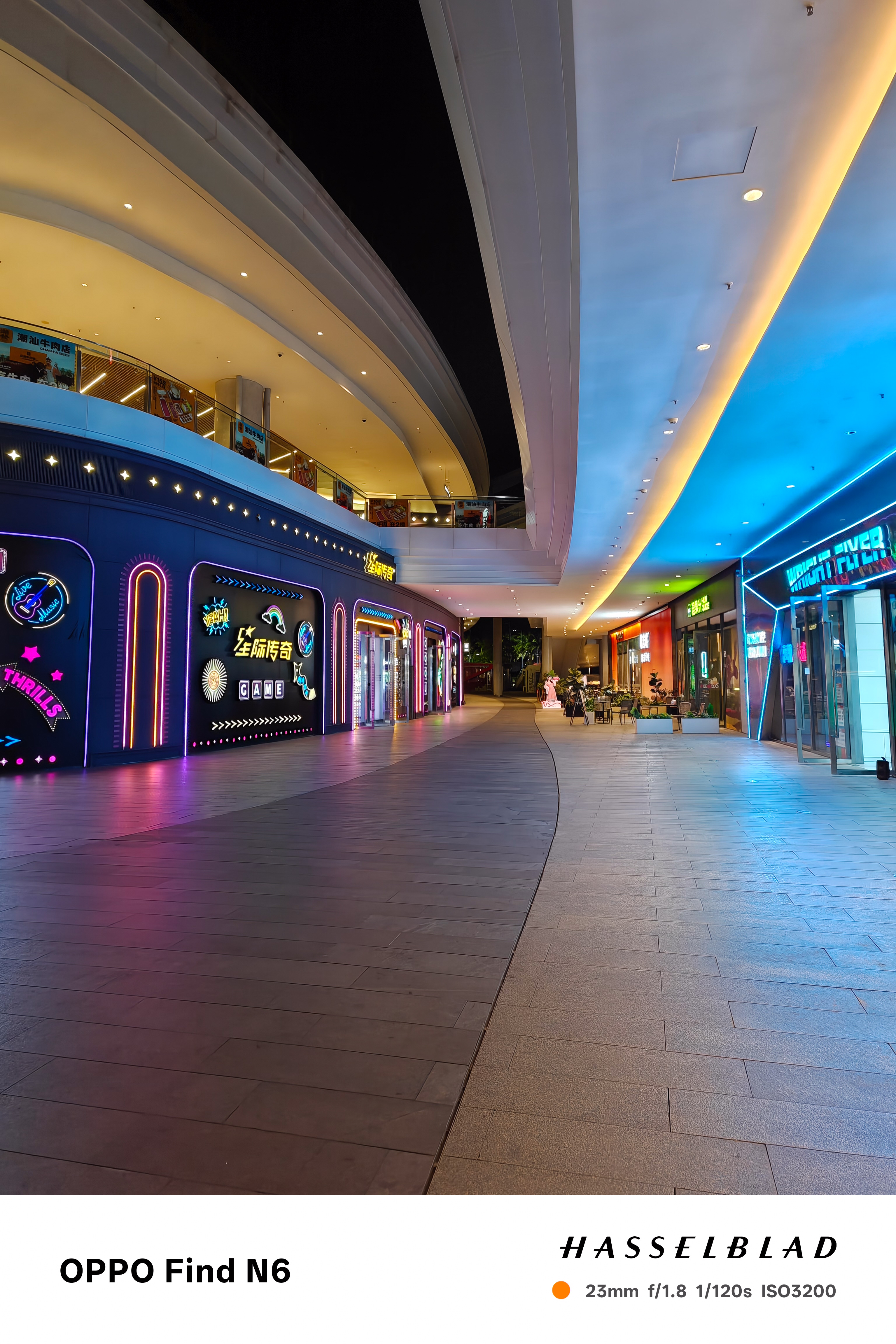 A wide-angle night shot of a futuristic outdoor shopping plaza. The walkway is illuminated by vibrant neon lights in shades of pink, blue, and yellow. The architecture features sweeping, curved white rooflines and glowing storefronts under a dark night sky.