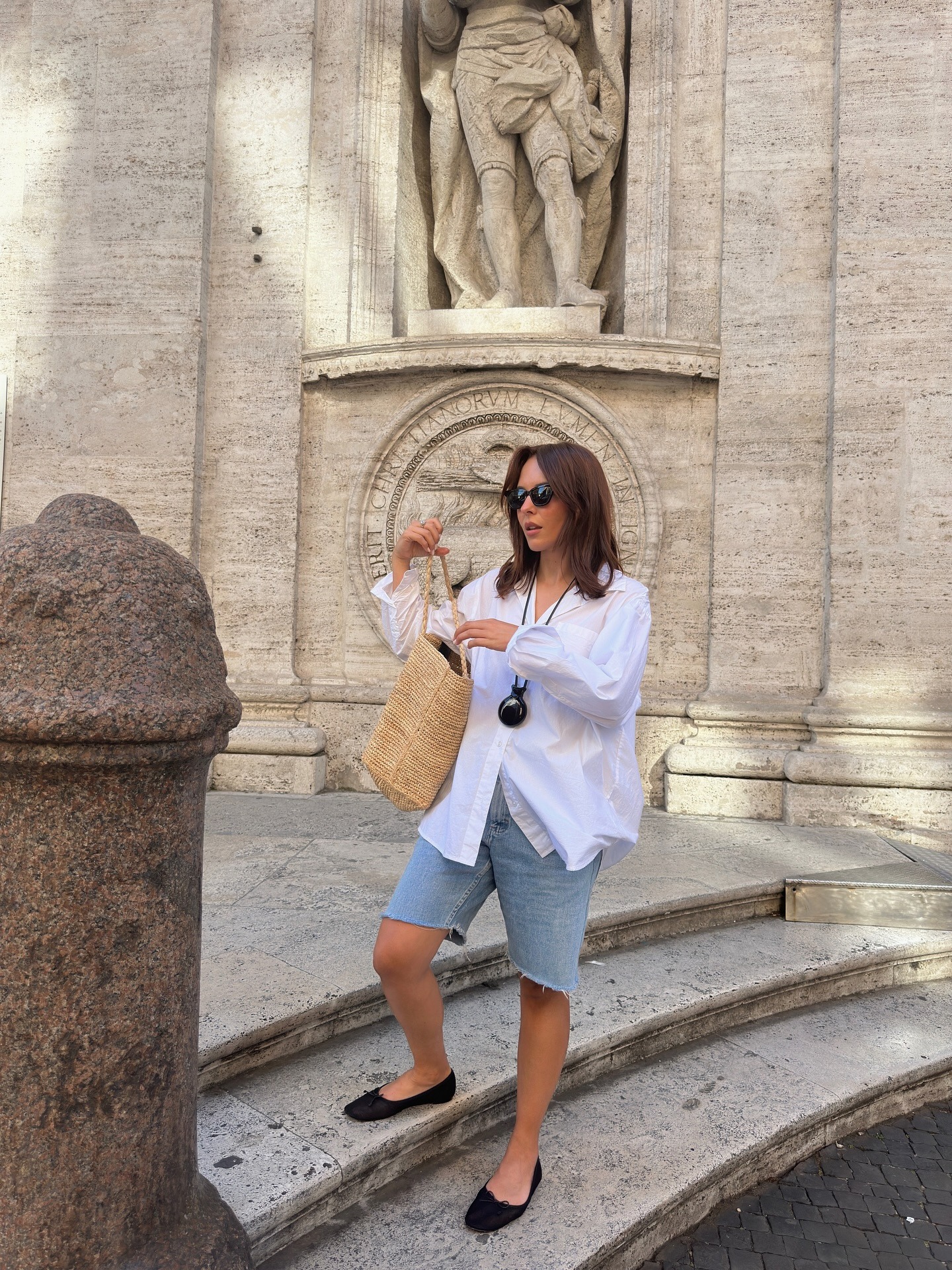 Influencer Debora wearing light jean shorts, a white button-up, and a woven straw bag.