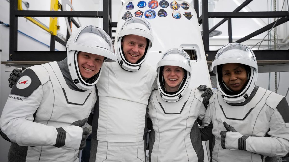 four people in white spacesuits with the visors up pose and smile in front of a white cone-shaped spacecraft