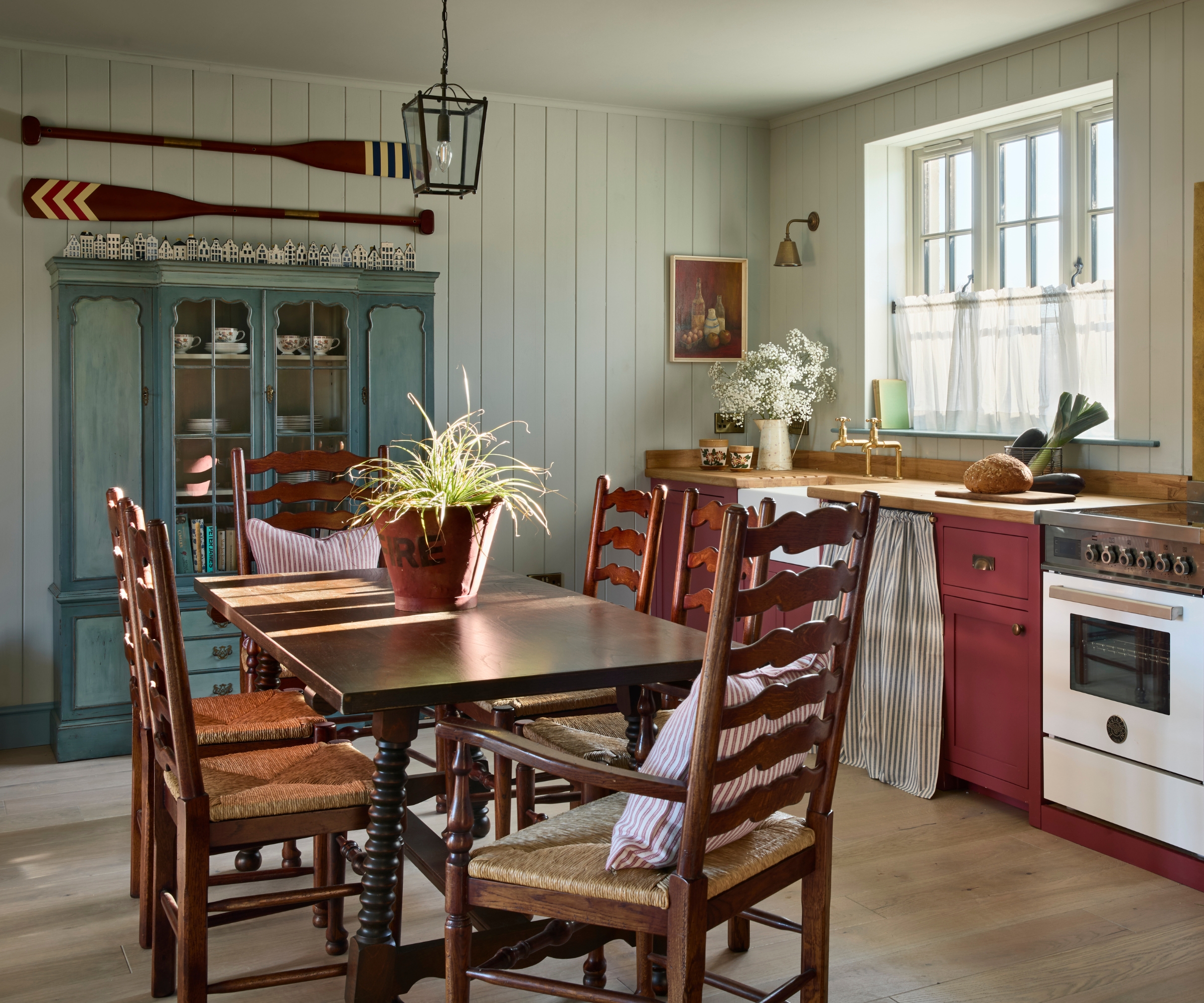 A coastal cottage kitchen with light blue paneled walls, wood countertops, burgundy cabinets, a dark wood kitchen table and chairs, and a freestanding antique cabinet painted mid blue