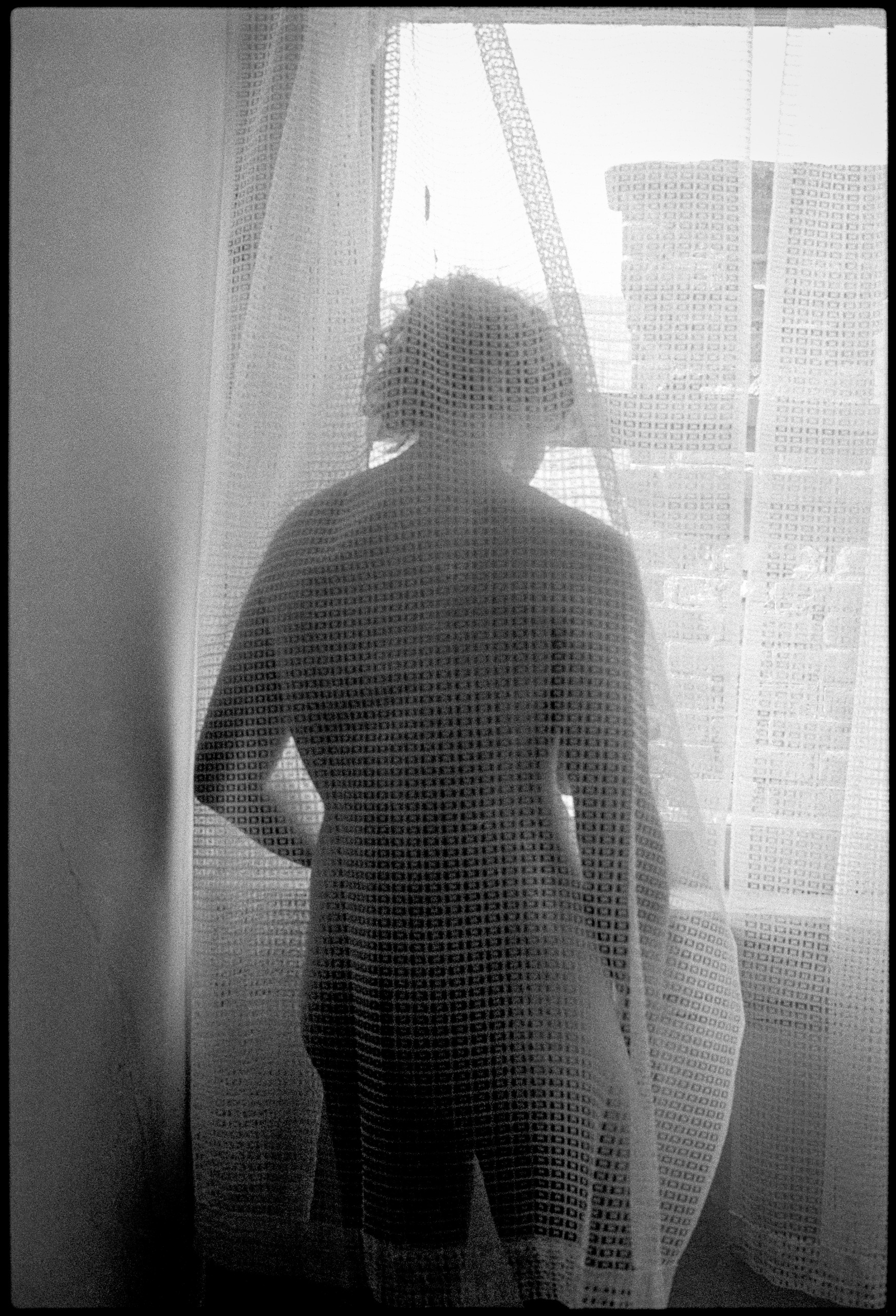 A black-and-white silhouette of a person standing behind a sheer patterned lace curtain in front of a window.
