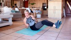 Woman exercising at home. She is sitting on an exercise mat with her feet raised, holding a weight plate and her torso turned to the right-hand side.