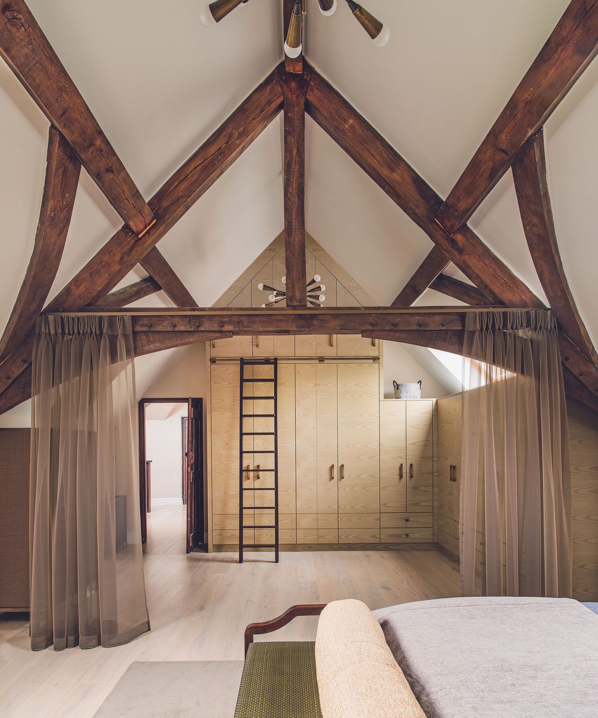Master bedroom with a wall of storage and sliding ladder to reach the top level, wardrobes and exposed beams under a vaulted ceiling, and net curtains drawn back.
