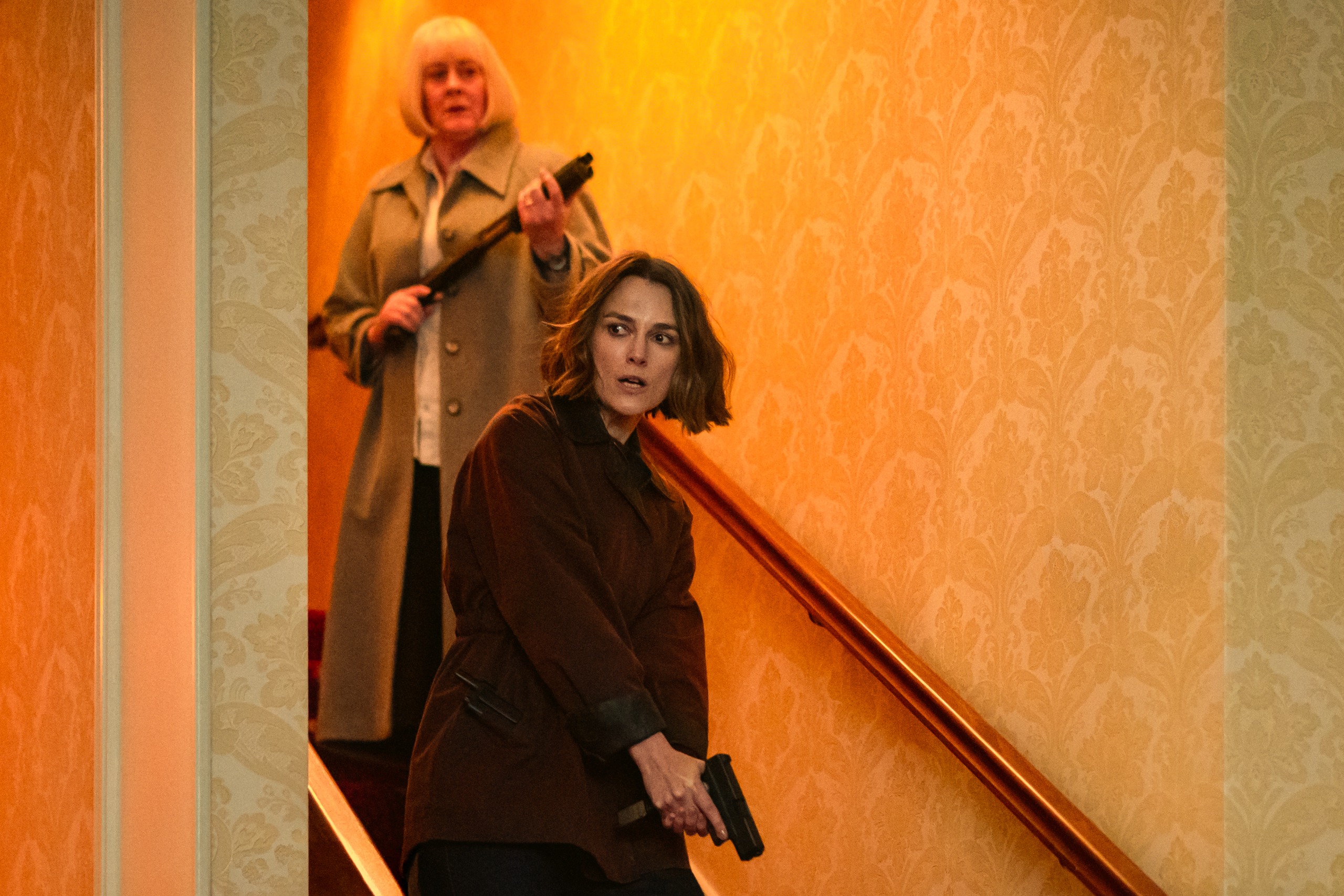 Sarah Lancashire and keira knightley wear coats and hold guns as they walk down a staircase in black doves season 2