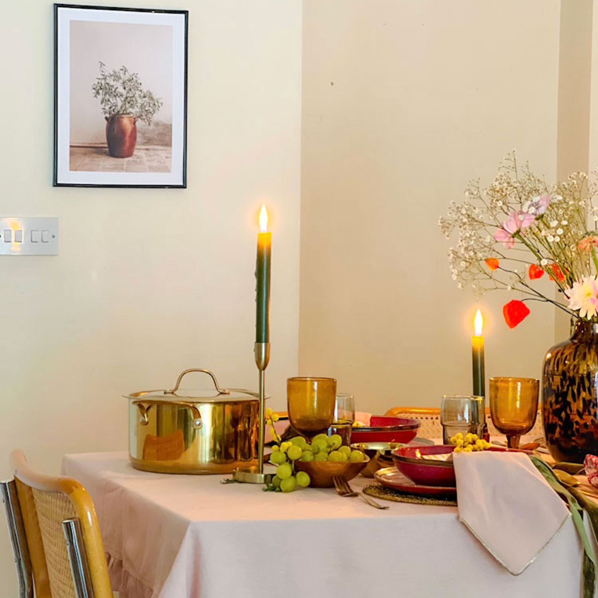 Table with tablecloth, metallic pot, golden candlesticks with lit pillar candles, amber wine glasses and tortoiseshell vase with fresh flowers