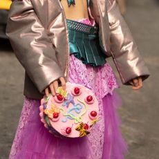 A street style image of a woman wearing a frilly pink skirt and holding a clutch bag shaped like a cake, reminiscent of the best gourmand perfumes