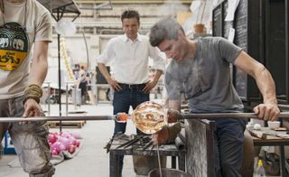 Workshop image, glass blower making a glass piece for the water feature, another man helping to hold a metal bar in place, man in the back ground hands on hips watching the process, industrial space blurred in the backdrop